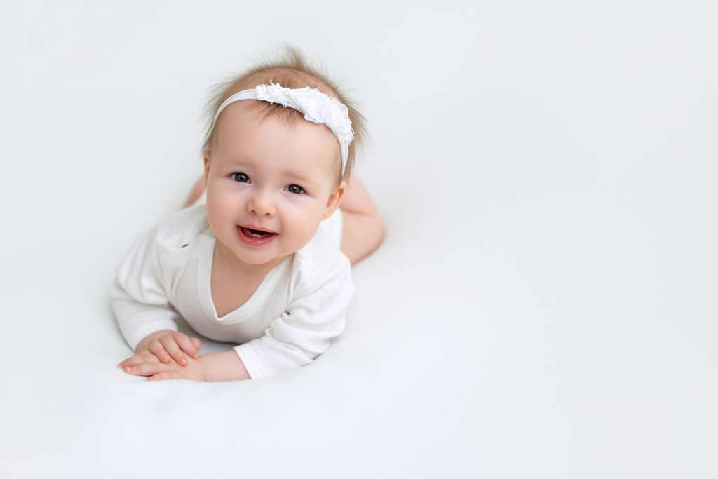 Bébé, vêtu d'un body blanc et d'un serre-tête, sourit gentiment, allongé sur le ventre.