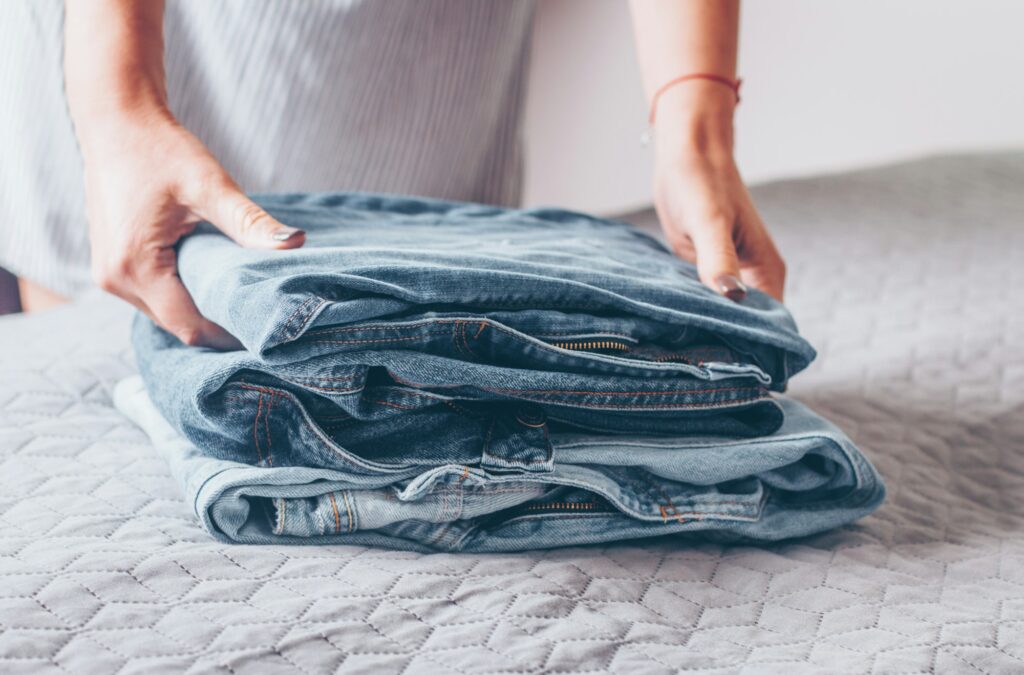 Woman folds a jeans on grey cover on bed. Pile of blue jeans clothes.