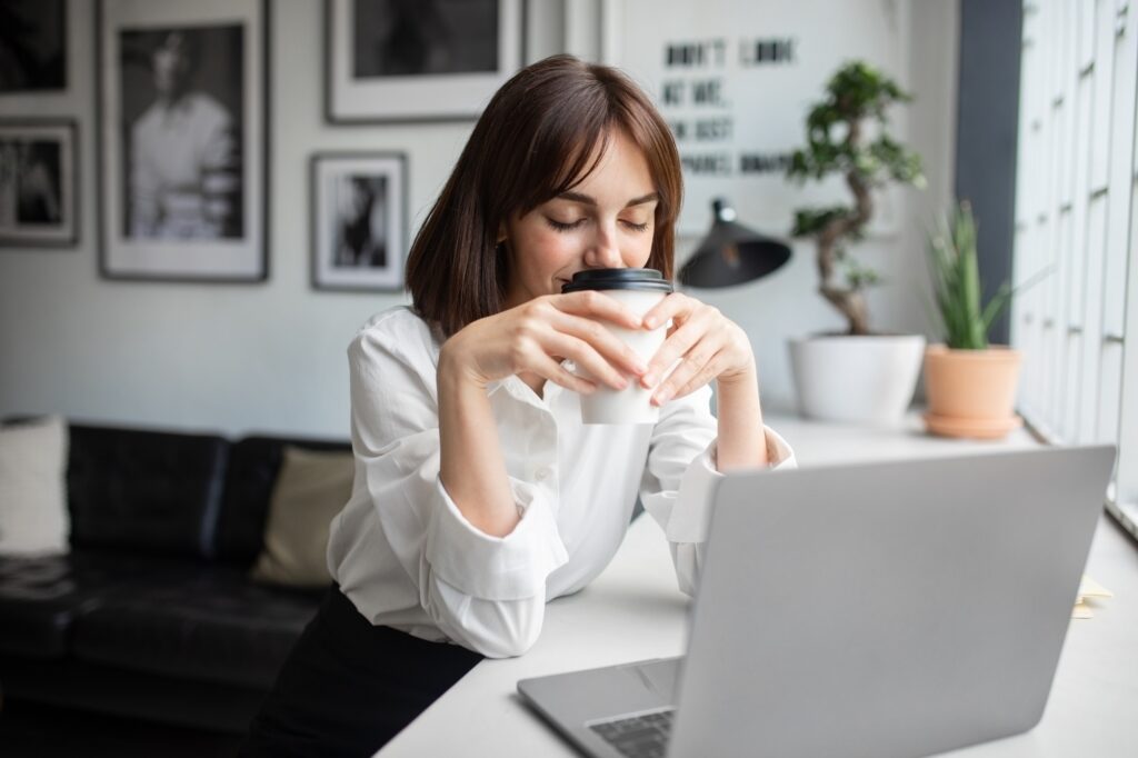 Happy young businesswoman drinking and enjoying coffee while having a break from work, sitting at